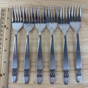 May include: Set of six stainless steel forks with a decorative design on the handles. The forks are arranged in a row on a wooden surface. Each fork has four tines and measures approximately 7 inches in length.