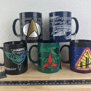 May include: Five ceramic mugs with Star Trek designs. The mugs are black, blue, and green, featuring logos and imagery from the series. One mug has the text "The Next Generation". The mugs are on a wooden surface.