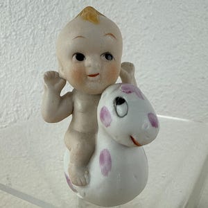 May include: A vintage porcelain figurine of a baby riding a white and pink spotted creature. The baby has a peach complexion, black eyes, and a small smile. The creature has a friendly expression.