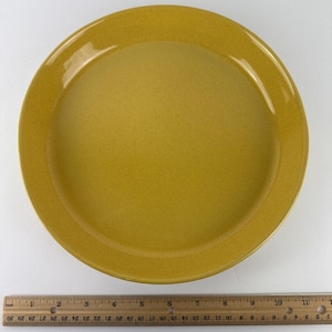 May include: A round, mustard-yellow plate with a speckled texture. The plate is approximately 11 inches in diameter. The plate has a slightly raised rim and a smooth, glossy finish. A wooden ruler is placed below the plate.