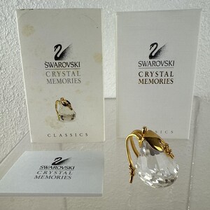 May include: A clear crystal pear-shaped figurine with a gold-colored stem and leaf accents. The figurine is displayed with its original packaging, which includes the text "SWAROVSKI CRYSTAL MEMORIES" and the Swarovski logo.