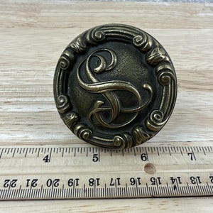 May include: Antique brass-toned drawer pull with an ornate design. The center features a stylized letter "S". The pull is round, approximately 2 inches in diameter, with a decorative border.