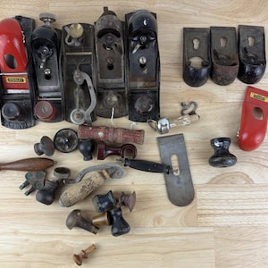 May include: A collection of vintage woodworking tools, including hand planes, handles, and other hardware. The tools are made of metal and wood, with various colors and finishes. The arrangement is on a light wood surface.