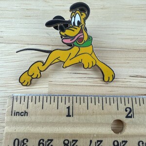 May include: A yellow Pluto pin with a black hat and green collar. The pin is approximately 2 inches long. Pluto is depicted in a running pose with his tongue sticking out. The pin is on a wooden surface.