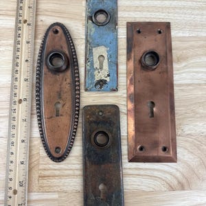 May include: An assortment of antique door hardware, including four different backplates and a ruler. The backplates vary in shape, color, and finish, with keyholes and knob holes. The ruler is marked in inches.