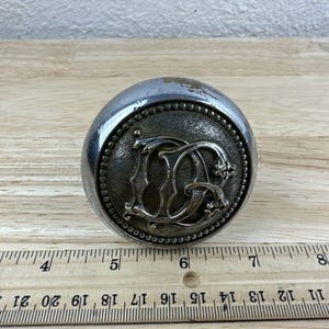 May include: A silver-toned door knob with a decorative, ornate design featuring the letters "DG" in the center.