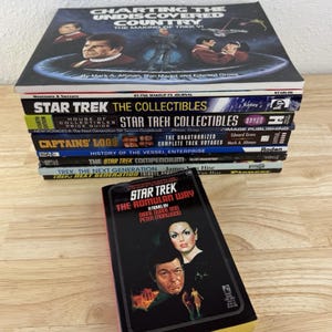 May include: A stack of Star Trek books, including "Charting the Undiscovered Country" and "Star Trek: The Romulan Way." The books feature images of characters and titles related to the Star Trek franchise. The books are stacked on a light-colored surface.