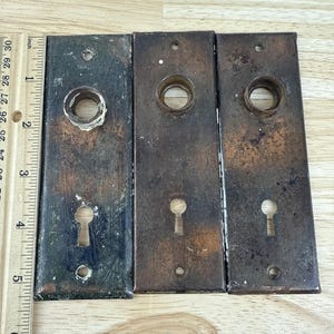 May include: Three antique metal door plates with keyhole openings. The plates are rectangular, with varying shades of brown and black, showing signs of age and wear. A wooden ruler is placed next to the plates.