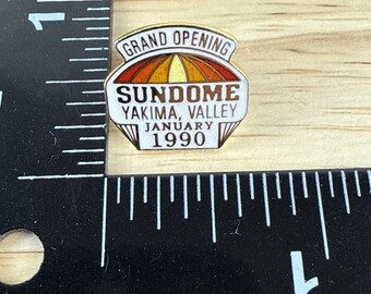 May include: A vintage pin with the text "GRAND OPENING" at the top, and "SUNDOME YAKIMA, VALLEY JANUARY 1990" below. The pin features a colorful design with red, orange, and yellow hues, set against a white background.