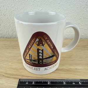 May include: White ceramic mug with a curved handle. The mug features a Starfleet Academy emblem with the text "Starfleet Academy, San Francisco, Ex Astra, Scientia, MMCLXI". The emblem includes a gold and brown design.