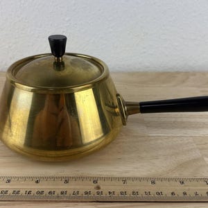 May include: A vintage brass saucepan with a black handle and lid knob. The pot has a shiny, golden finish and is sitting on a wooden surface. A ruler is placed next to the pot, showing inch measurements.