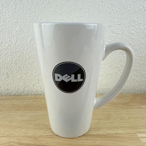 May include: A white ceramic mug with a black handle and the Dell logo in a black circle. The mug is on a wooden surface with a ruler.