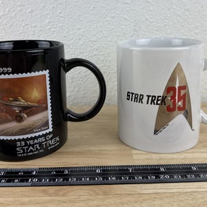 May include: Two ceramic Star Trek mugs. One black mug features a stamp design with a spaceship and the text "33 Years of Star Trek." The other is white with the Star Trek logo and "35."