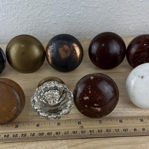 May include: A collection of antique door knobs in various styles and materials. The knobs include a dark blue marble, brass, and wood. A ruler is placed below the knobs, showing inches.