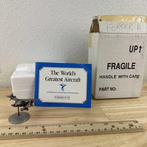 May include: A miniature metal Fokker D VII aircraft model on a round base, next to a blue card that reads "The World's Greatest Aircraft". A cardboard box labeled "Fokker A" and "Fragile" is also present. A wooden ruler is in the foreground.