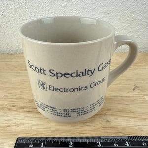 May include: A beige ceramic mug with a handle, featuring the dark blue text Scott Specialty Gases Electronics Group and a logo with the letters SGP. A black ruler is positioned at the base, showing the mug is approximately 4 inches wide.