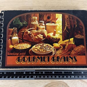 May include: A vintage cookbook titled "Cooking with Gourmet Grains" with a black spiral binding. The cover features a rustic illustration of various baked goods, grains, and a pumpkin. The text is in a decorative font.