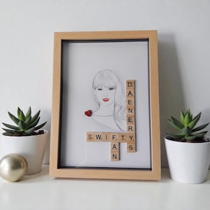 Peut inclure: Un croquis au crayon encadré d'une femme au rouge à lèvres rouge et à un cœur rouge. Le cadre comprend des lettres en bois épelant "DAENER" et "SWIFTY AN". Deux plantes succulentes en pot et une sphère dorée sont sur la surface blanche.