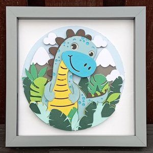 May include: A framed paper craft featuring a blue dinosaur with yellow stripes and brown spots, surrounded by green leaves and white mountains. The background is light blue.