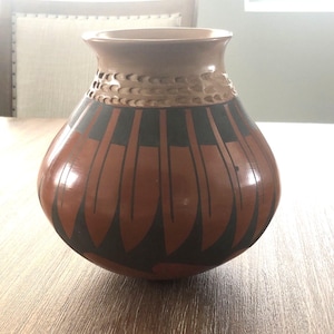 May include: A rounded ceramic vase with a smooth, light tan neck featuring a textured, scalloped band. The body of the vase is decorated with a repeating pattern of vertical, elongated black and terracotta-colored leaf-like shapes.