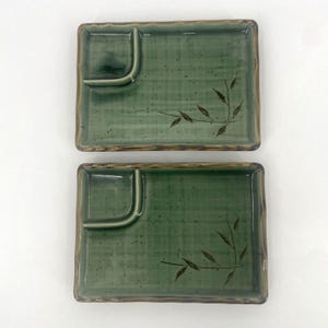 May include: Two rectangular ceramic plates in a sage green glaze with a dark brown bamboo leaf motif. Each plate features a raised, curved corner partition, a subtle grid pattern, and a dark brown rim, creating a functional surface for serving.