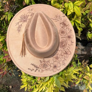 May include: A tan fedora hat with a teardrop crown and a wide brim. The brim is decorated with floral designs and a brown leather band. A tassel hangs from the side of the hat. The hat is set against a background of green and yellow foliage.