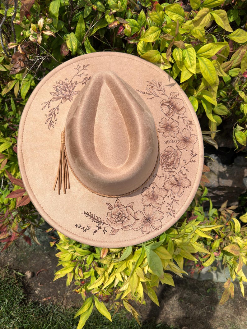May include: A tan fedora hat with a teardrop crown and a wide brim. The brim is decorated with floral designs and a brown leather band. A tassel hangs from the side of the hat. The hat is set against a background of green and yellow foliage.
