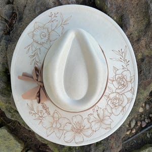 May include: A cream-coloured fedora hat with a brown ribbon and floral design. The hat features an engraved pattern of flowers and leaves around the brim. The hat's crown is a lighter cream colour.