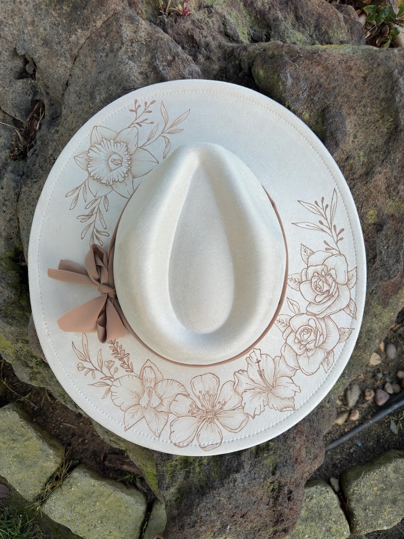 May include: A cream-coloured fedora hat with a brown ribbon and floral design. The hat features an engraved pattern of flowers and leaves around the brim. The hat's crown is a lighter cream colour.