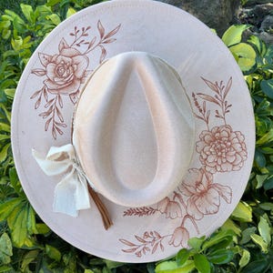 May include: A light beige fedora hat with a wide brim, decorated with floral patterns in a darker beige. A cream ribbon and a brown leather cord are attached. The hat is displayed against a background of green leaves.