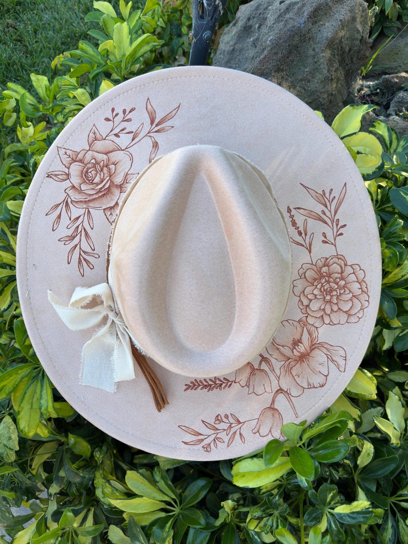May include: A light beige fedora hat with a wide brim, decorated with floral patterns in a darker beige. A cream ribbon and a brown leather cord are attached. The hat is displayed against a background of green leaves.