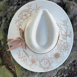 May include: A cream-coloured fedora hat with a wide brim, decorated with floral line art in shades of brown. A brown ribbon is tied around the base of the crown. The hat is resting on a textured stone surface.