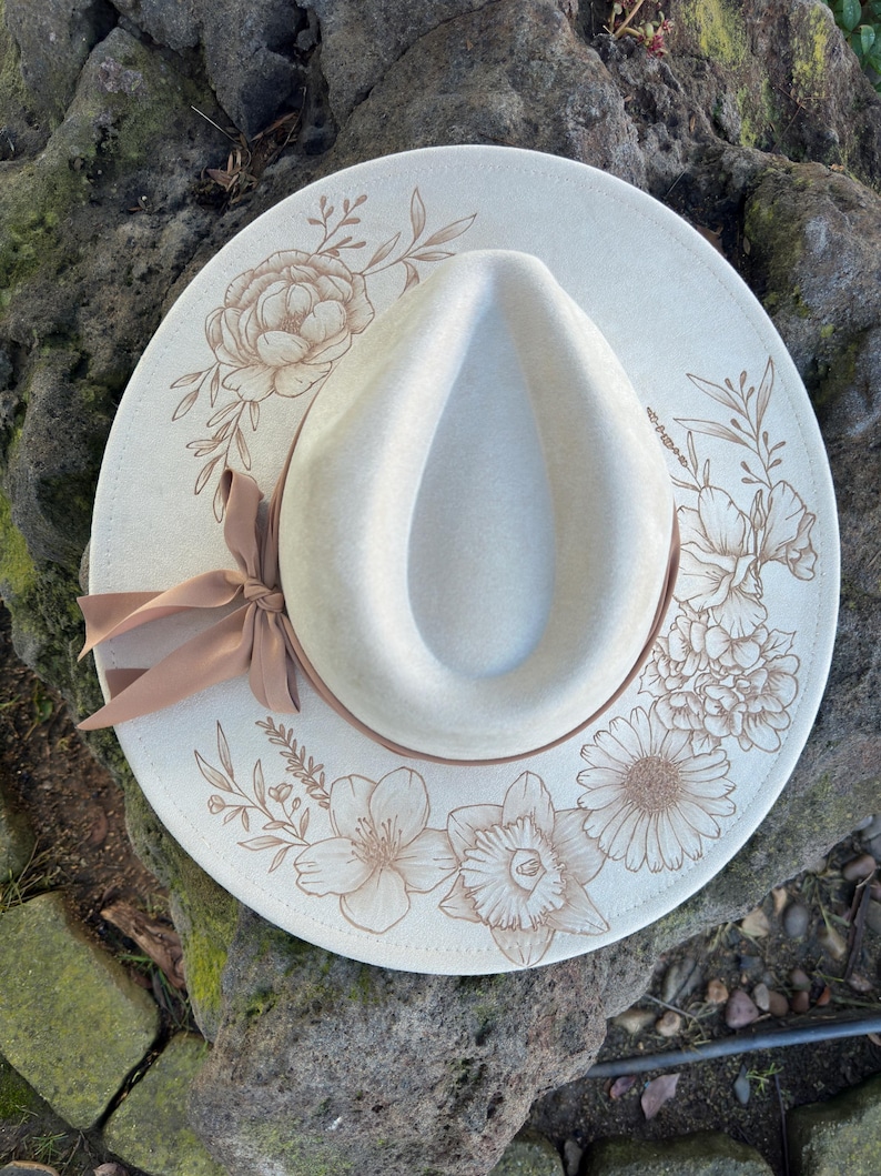 May include: A cream-coloured fedora hat with a wide brim, decorated with floral line art in shades of brown. A brown ribbon is tied around the base of the crown. The hat is resting on a textured stone surface.
