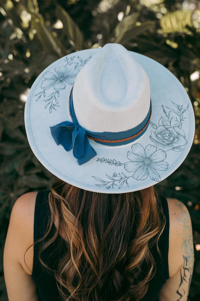 May include: A light blue fedora hat with a white crown and a wide brim. The brim features floral illustrations in a darker blue. A dark blue ribbon and a brown band encircle the hat.