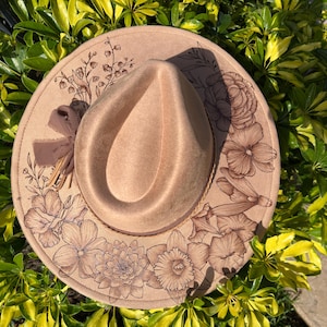 May include: A beige fedora hat with an etched floral design around the brim. The hat has a brown ribbon and a detailed pattern of flowers and leaves. The hat is set against a backdrop of green and yellow foliage.