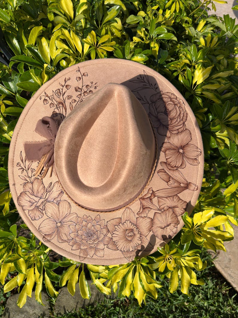 May include: A beige fedora hat with an etched floral design around the brim. The hat has a brown ribbon and a detailed pattern of flowers and leaves. The hat is set against a backdrop of green and yellow foliage.