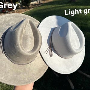 May include: Two fedora hats, one grey and one light grey. Each hat features a wide brim, a pinched crown, and a decorative band with tassels. The hats are made from a soft, suede-like material, suitable for various occasions.