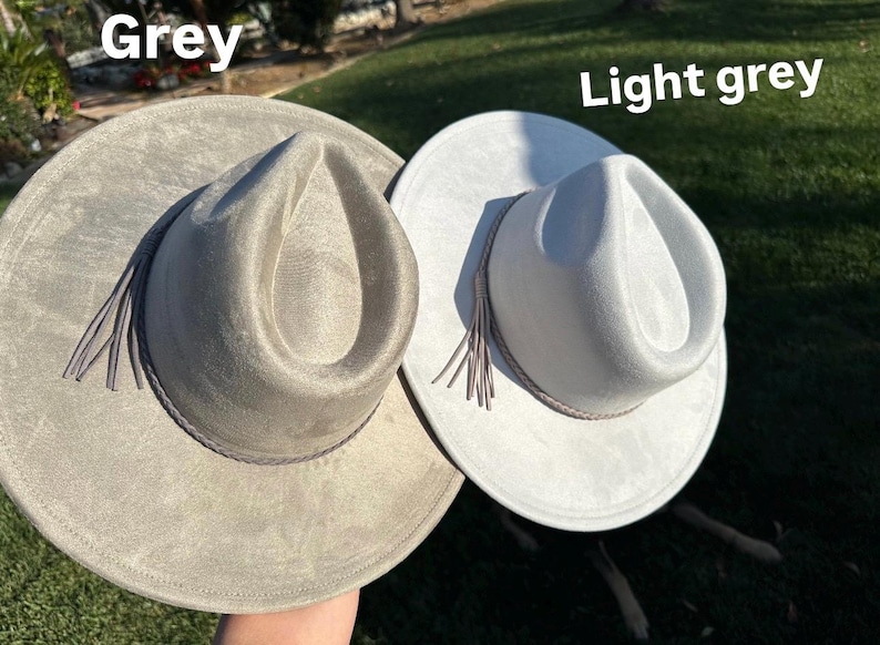 May include: Two fedora hats, one grey and one light grey. Each hat features a wide brim, a pinched crown, and a decorative band with tassels. The hats are made from a soft, suede-like material, suitable for various occasions.