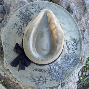 May include: A sage green fedora hat with a wide brim. The brim features blue floral designs and a black ribbon bow. The hat is displayed on a gray stone surface.