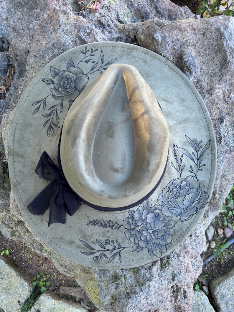 May include: A sage green fedora hat with a wide brim. The brim features blue floral designs and a black ribbon bow. The hat is displayed on a gray stone surface.