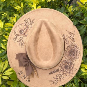 May include: A tan fedora hat with etched floral and leaf designs around the brim. A brown ribbon and feather adorn the hat's band. The date "08.29.26" is printed on the brim. The hat is surrounded by green foliage.