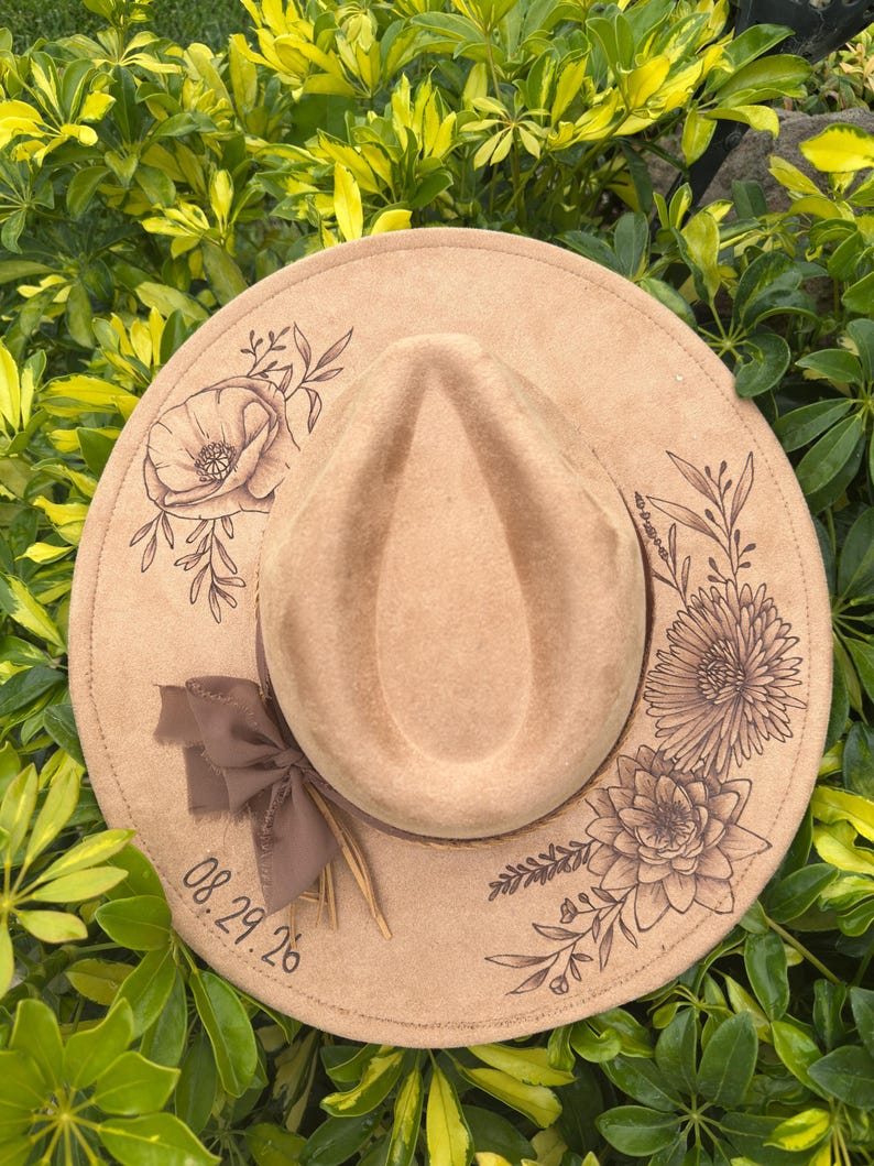 May include: A tan fedora hat with etched floral and leaf designs around the brim. A brown ribbon and feather adorn the hat's band. The date "08.29.26" is printed on the brim. The hat is surrounded by green foliage.