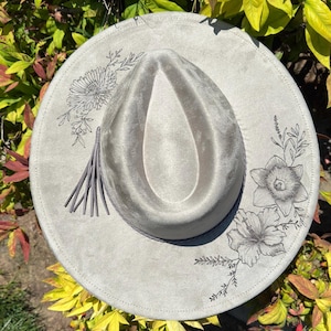 May include: A light grey fedora hat with a wide brim. The hat has a decorative floral design in black ink around the brim and a grey tassel. The hat is surrounded by green and yellow foliage.
