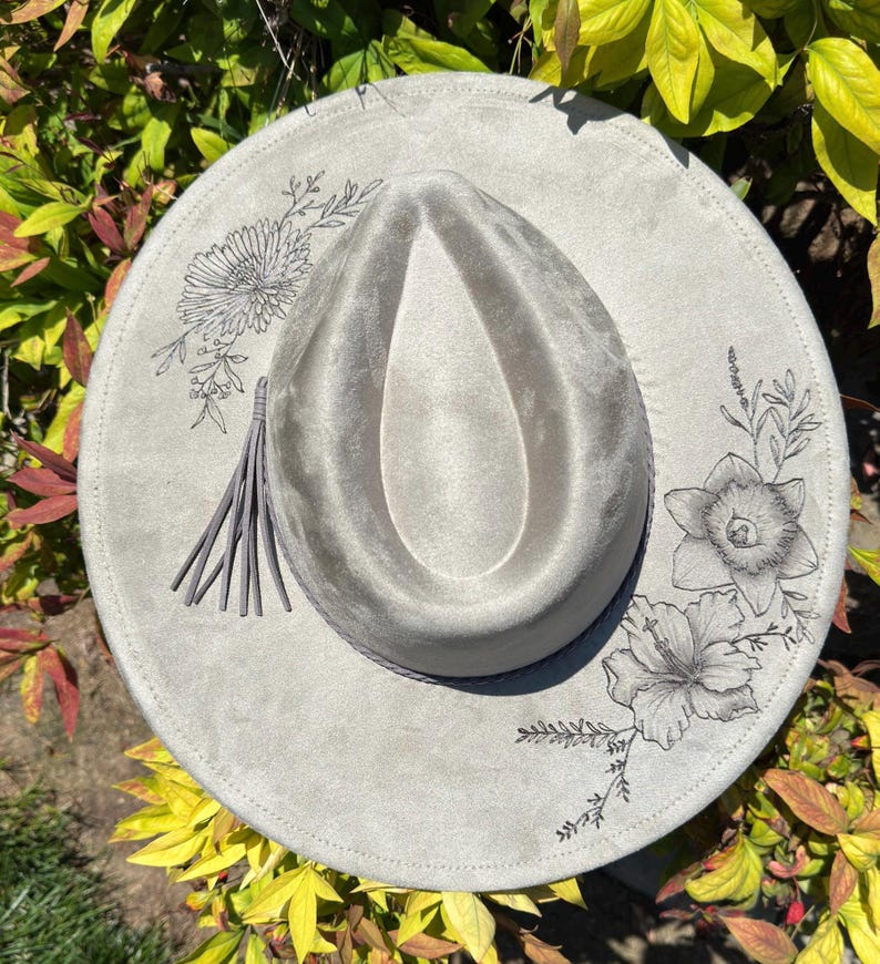 May include: A light grey fedora hat with a wide brim. The hat has a decorative floral design in black ink around the brim and a grey tassel. The hat is surrounded by green and yellow foliage.