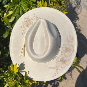 May include: A cream-coloured fedora hat with a wide brim. The hat features floral embroidery in a light brown colour on the brim. A decorative tassel hangs from the side. The hat is surrounded by green foliage.
