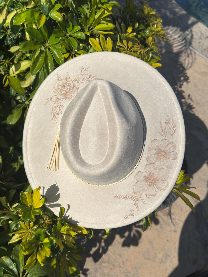 May include: A cream-coloured fedora hat with a wide brim. The hat features floral embroidery in a light brown colour on the brim. A decorative tassel hangs from the side. The hat is surrounded by green foliage.