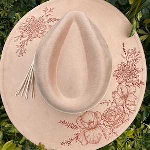 May include: A light pink fedora hat with a wide brim. The hat has a floral design in a darker pink around the brim. A tassel hangs from the side. The hat is set against a backdrop of green foliage.