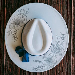 May include: A pale blue fedora hat with a white crown and brim. The brim features floral line art and a navy blue ribbon. The hat is set against a dark wood grain background.
