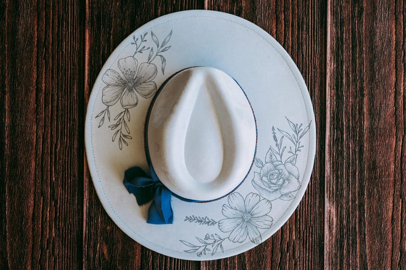 May include: A pale blue fedora hat with a white crown and brim. The brim features floral line art and a navy blue ribbon. The hat is set against a dark wood grain background.