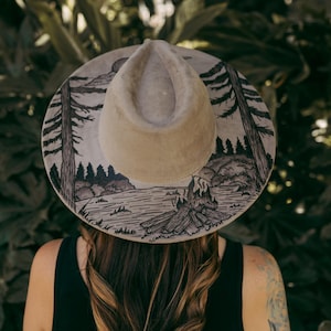 May include: A beige fedora hat with a detailed black and white landscape design around the brim. The artwork depicts a forest, lake, and campfire scene. The hat has a classic teardrop crown.